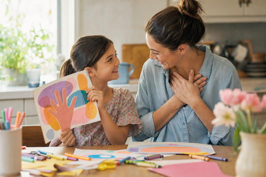 Child showing their mother a colorful handprint keepsake at a sunlit kitchen craft table