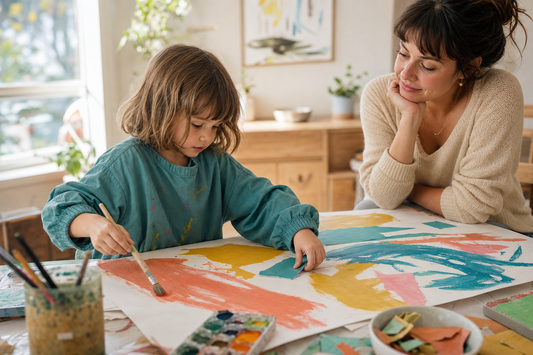 Preschooler painting and arranging torn paper at a sunny art table while an adult watches nearby
